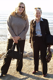 Two people standing together outdoors on a beach with water behind them.